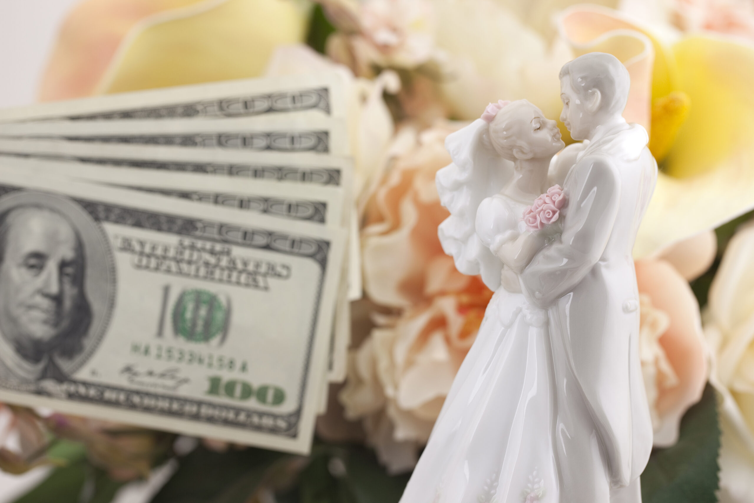 A wedding cake topper showing a groom and bride with one hundred dollars fanned out in the background with flowers